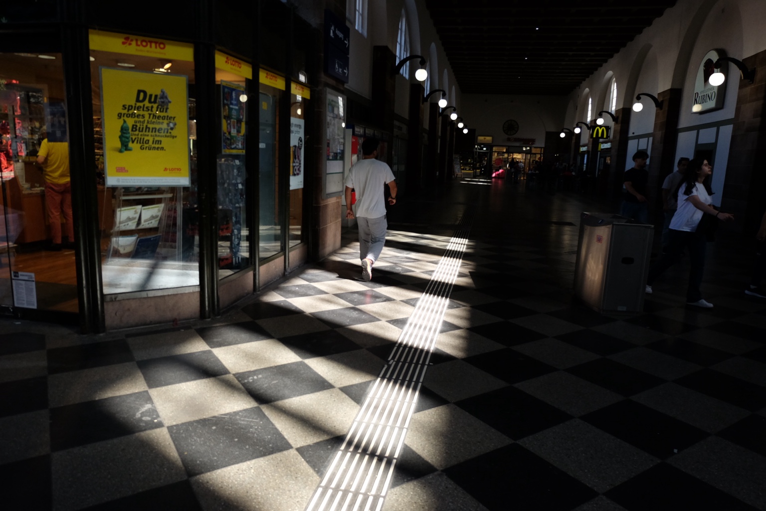 Person walking through a station hall with checkerboard floor and strong light