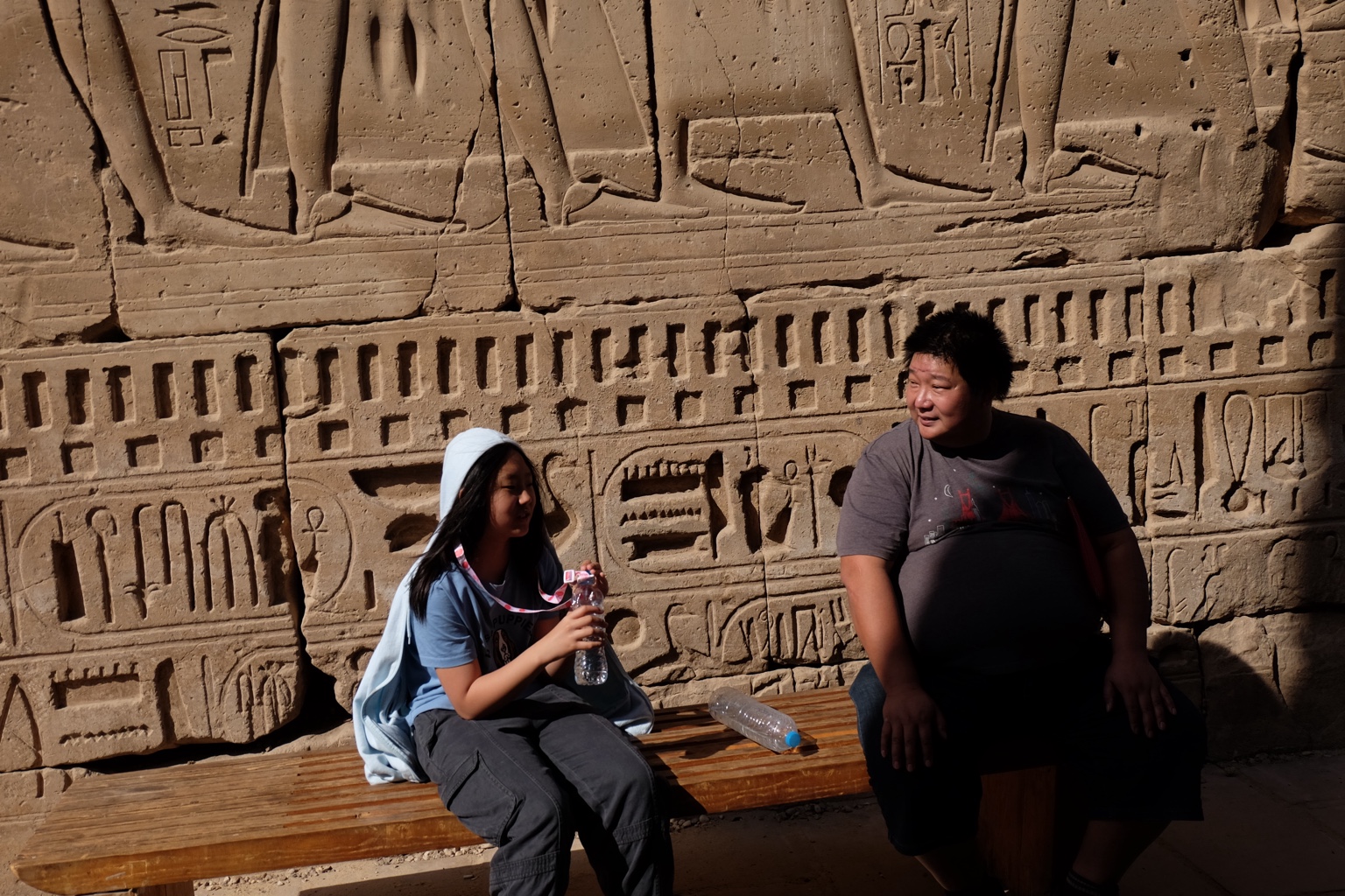 Two people resting on a bench before a carved stone wall