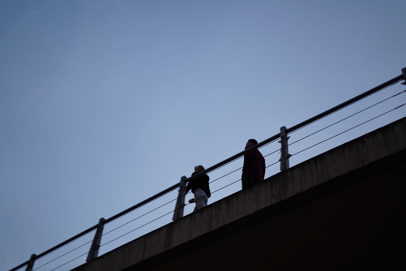 Two silhouetted people walking along a bridge against a blue sky