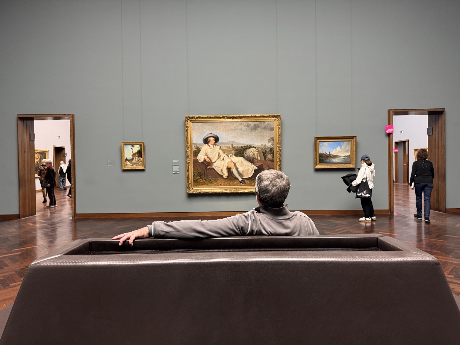 A person seated on a museum bench looking toward framed paintings