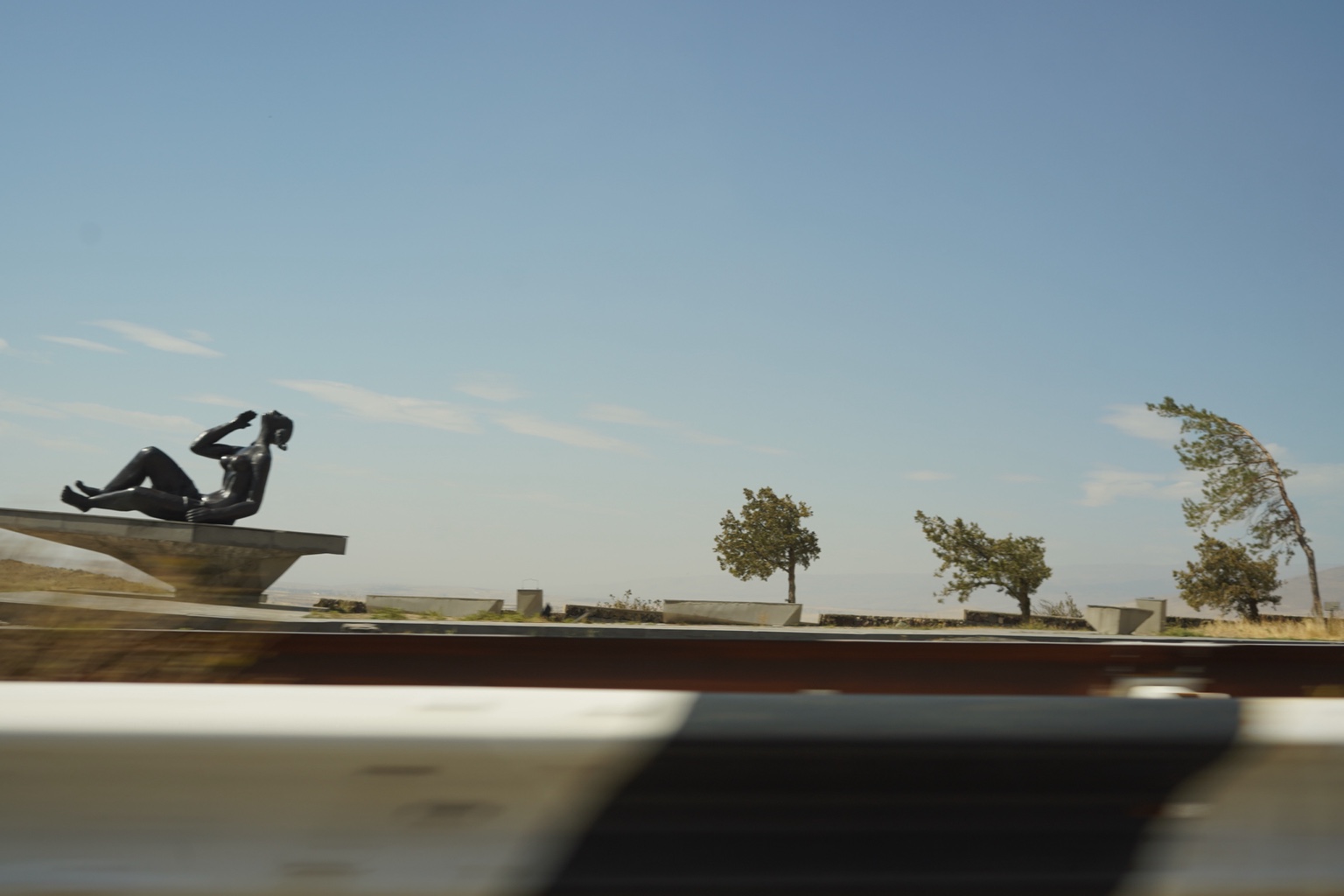 Roadside statue and wind-bent trees seen through a moving vehicle